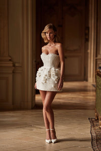 A dazzling bustier mini dress embellished with sequins, pearls, and 3D flowers by hand. Featuring a heart-shaped neckline with accentuated built-in cups, a cinched waist, and a structured tulip skirt adorned with 3D tulle flowers that add a sculpted finish. The look can be elevated with tulle long gloves adorned with volumetric flower appliqués. Perfect as a second wedding dress or a go-out party dress.