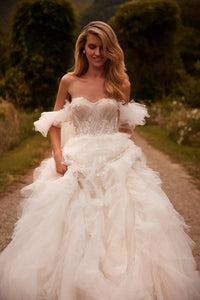 Introducing Dress Esmeralda - a stunning, voluminous dress in a light nude tone with a detachable train. The off-the-shoulder corset is adorned with delicate lace and handmade embroidery, while the flowing sleeves and ruffled bodice add a touch of elegance. The airy skirt is adorned with intricate lace and 3D floral appliques, creating a truly unique and unforgettable look. Perfect for any special occasion.