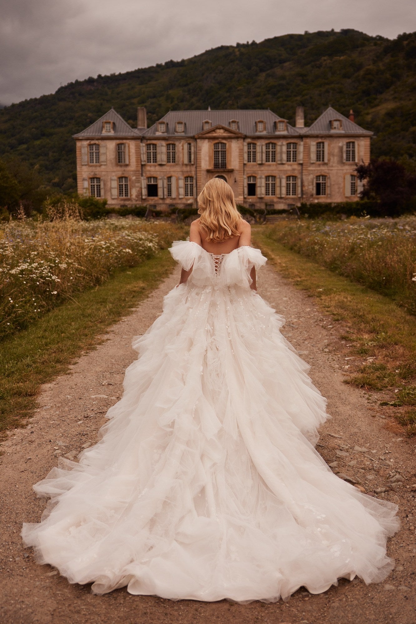 Introducing Dress Esmeralda - a stunning, voluminous dress in a light nude tone with a detachable train. The off-the-shoulder corset is adorned with delicate lace and handmade embroidery, while the flowing sleeves and ruffled bodice add a touch of elegance. The airy skirt is adorned with intricate lace and 3D floral appliques, creating a truly unique and unforgettable look. Perfect for any special occasion.