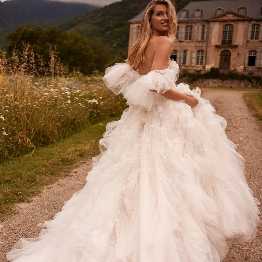 Introducing Dress Esmeralda - a stunning, voluminous dress in a light nude tone with a detachable train. The off-the-shoulder corset is adorned with delicate lace and handmade embroidery, while the flowing sleeves and ruffled bodice add a touch of elegance. The airy skirt is adorned with intricate lace and 3D floral appliques, creating a truly unique and unforgettable look. Perfect for any special occasion.