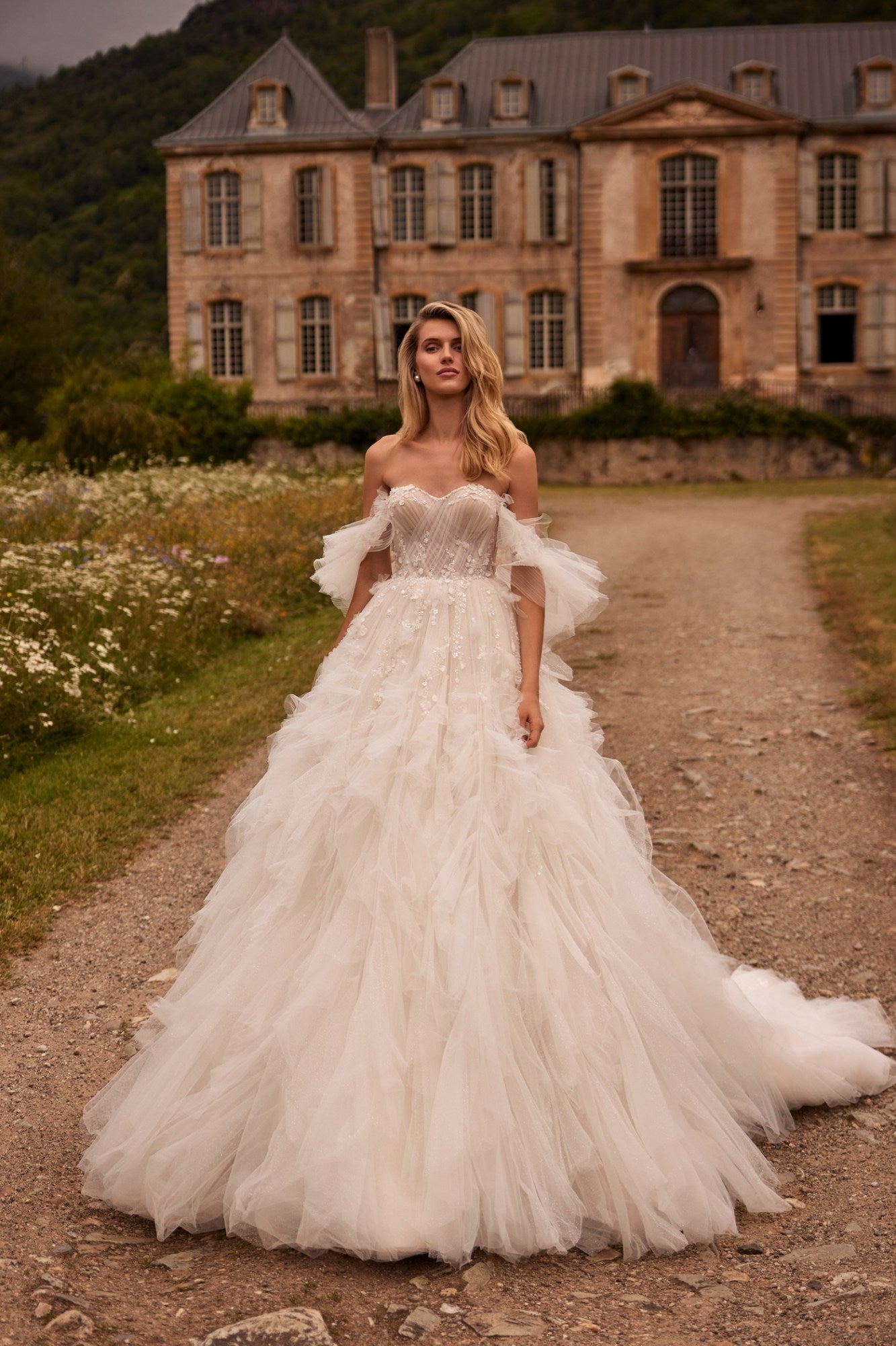 Introducing Dress Esmeralda - a stunning, voluminous dress in a light nude tone with a detachable train. The off-the-shoulder corset is adorned with delicate lace and handmade embroidery, while the flowing sleeves and ruffled bodice add a touch of elegance. The airy skirt is adorned with intricate lace and 3D floral appliques, creating a truly unique and unforgettable look. Perfect for any special occasion.