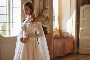 Experience ethereal elegance in the Anastasia Gown. Hand-embroidered with delicate lace, sequins, beads, pearls, 3D flowers, and butterflies, this nude a-line dress features an off-shoulder bodice with long sleeves and a hidden zipper closure. The airy mesh inserts on the sleeves and pleated skirt add a whimsical touch.