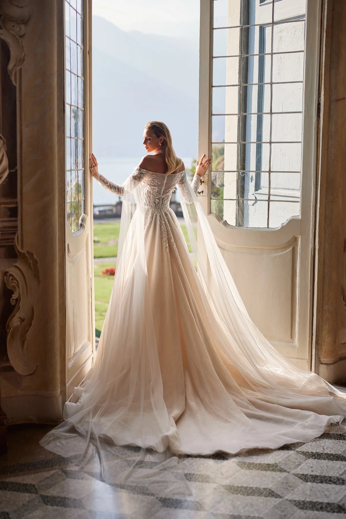 Experience ethereal elegance in the Anastasia Gown. Hand-embroidered with delicate lace, sequins, beads, pearls, 3D flowers, and butterflies, this nude a-line dress features an off-shoulder bodice with long sleeves and a hidden zipper closure. The airy mesh inserts on the sleeves and pleated skirt add a whimsical touch.
