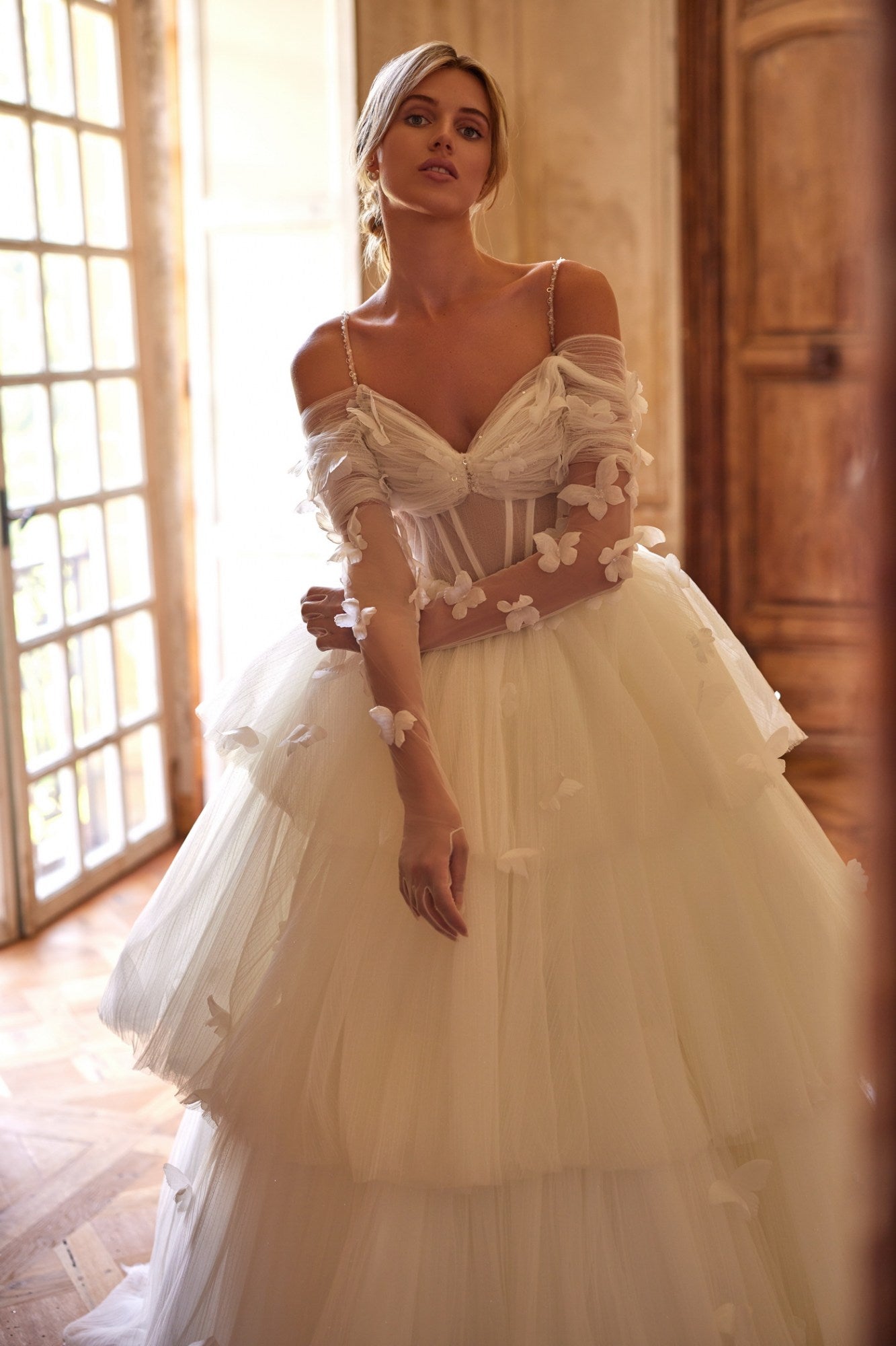 Experience a fairytale in our butterfly dress! Handmade 3D organza butterflies adorn the delicate pleated mesh, while a removable cup corset and embroidered skirt add romance. With off-shoulder straps and a hidden zipper, it's a playful take on the classic ball gown. Complete the look with embellished mesh gloves and a veil, both adorned with handmade butterfly and embroidery details.