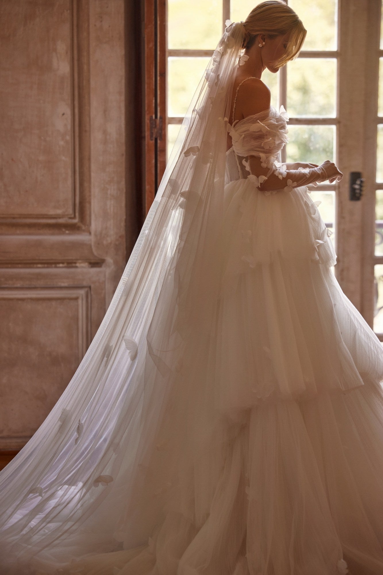 Experience a fairytale in our butterfly dress! Handmade 3D organza butterflies adorn the delicate pleated mesh, while a removable cup corset and embroidered skirt add romance. With off-shoulder straps and a hidden zipper, it's a playful take on the classic ball gown. Complete the look with embellished mesh gloves and a veil, both adorned with handmade butterfly and embroidery details.