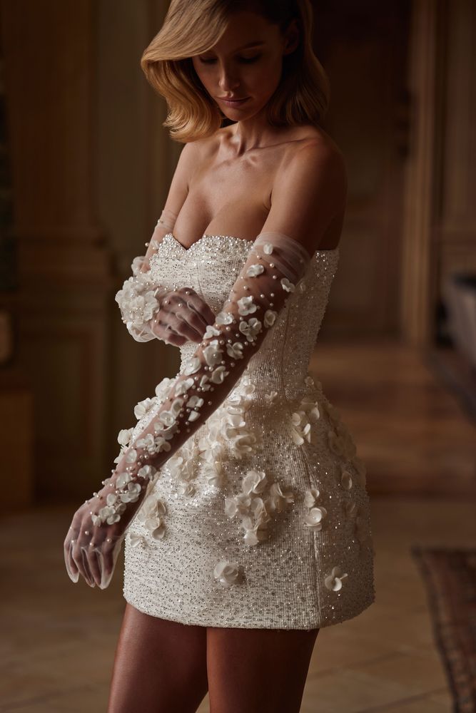 A dazzling bustier mini dress embellished with sequins, pearls, and 3D flowers by hand. Featuring a heart-shaped neckline with accentuated built-in cups, a cinched waist, and a structured tulip skirt adorned with 3D tulle flowers that add a sculpted finish. The look can be elevated with tulle long gloves adorned with volumetric flower appliqués. Perfect as a second wedding dress or a go-out party dress.
