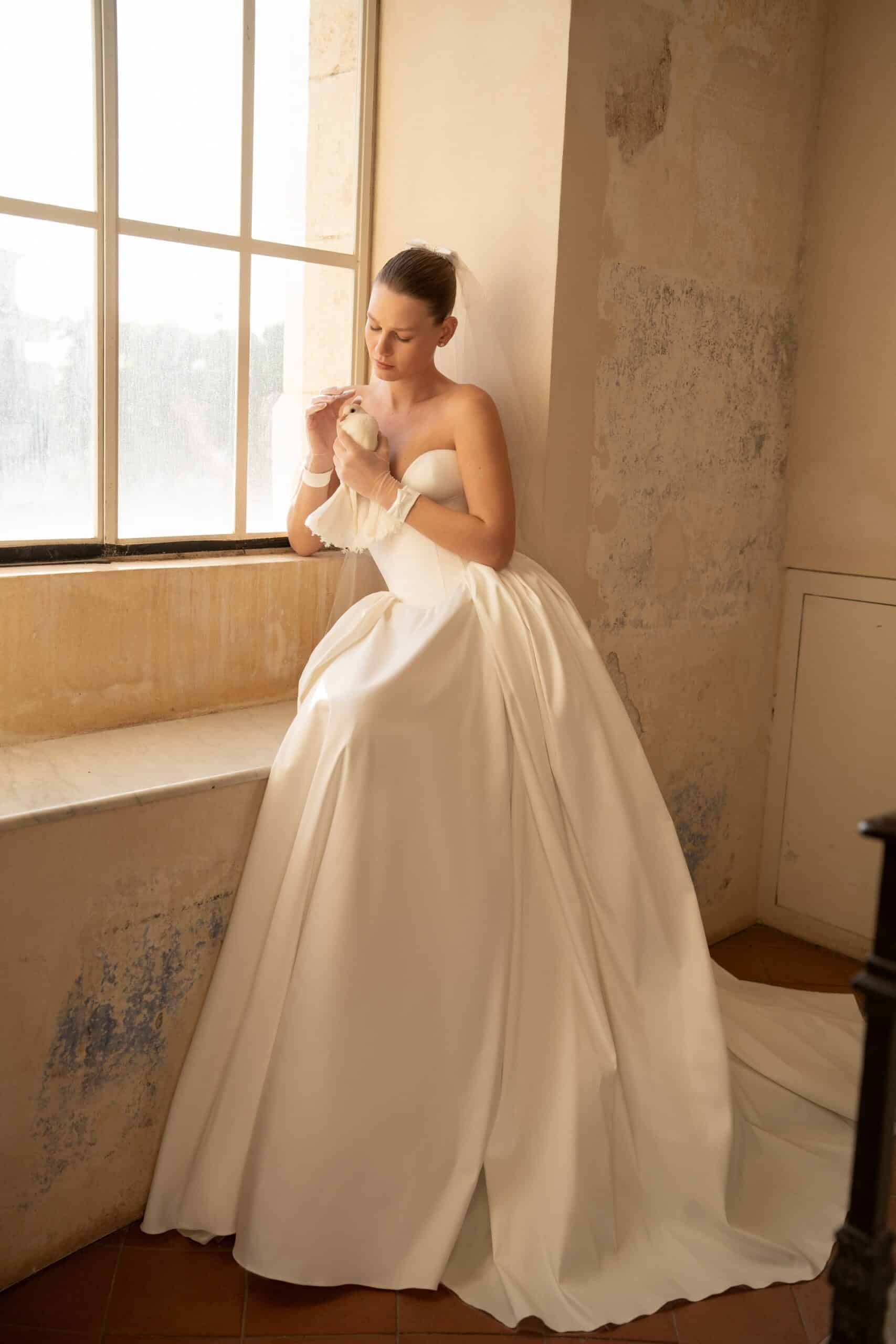 Elevate your special day with the Blue Gown. Made from luxurious Silk Mikado, it features a strapless sweetheart corset and basque waist for a flattering fit. The unique bow-shaped off-shoulder sleeves, coordinating gloves, and matching veil add a touch of elegance and sophistication. Create unforgettable memories in this stunning princess gown.
