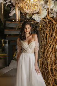 Elevate your special day with our Robe Mae. The elegant column silhouette features stunning beaded lace for a touch of glamour, while the off-shoulder sheer sleeves and deep v-neckline add a hint of romance. Complete the look with the delicate tulle skirt, for a dreamy and unforgettable bridal ensemble.