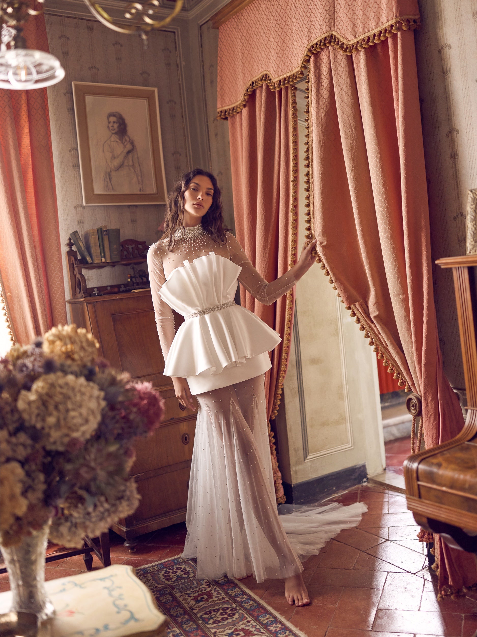 The Cassiopeia Gown captivates with its extraordinary seashell-inspired silhouette and radiant shimmer. Crafted from glossy mikado and crystal-embellished tulle, it features a neckline richly detailed with crystals and pearls for a sophisticated focal point. The bodice secures with hooks and a hidden snap at the waist, complemented by a matching embellished belt that defines the figure. 