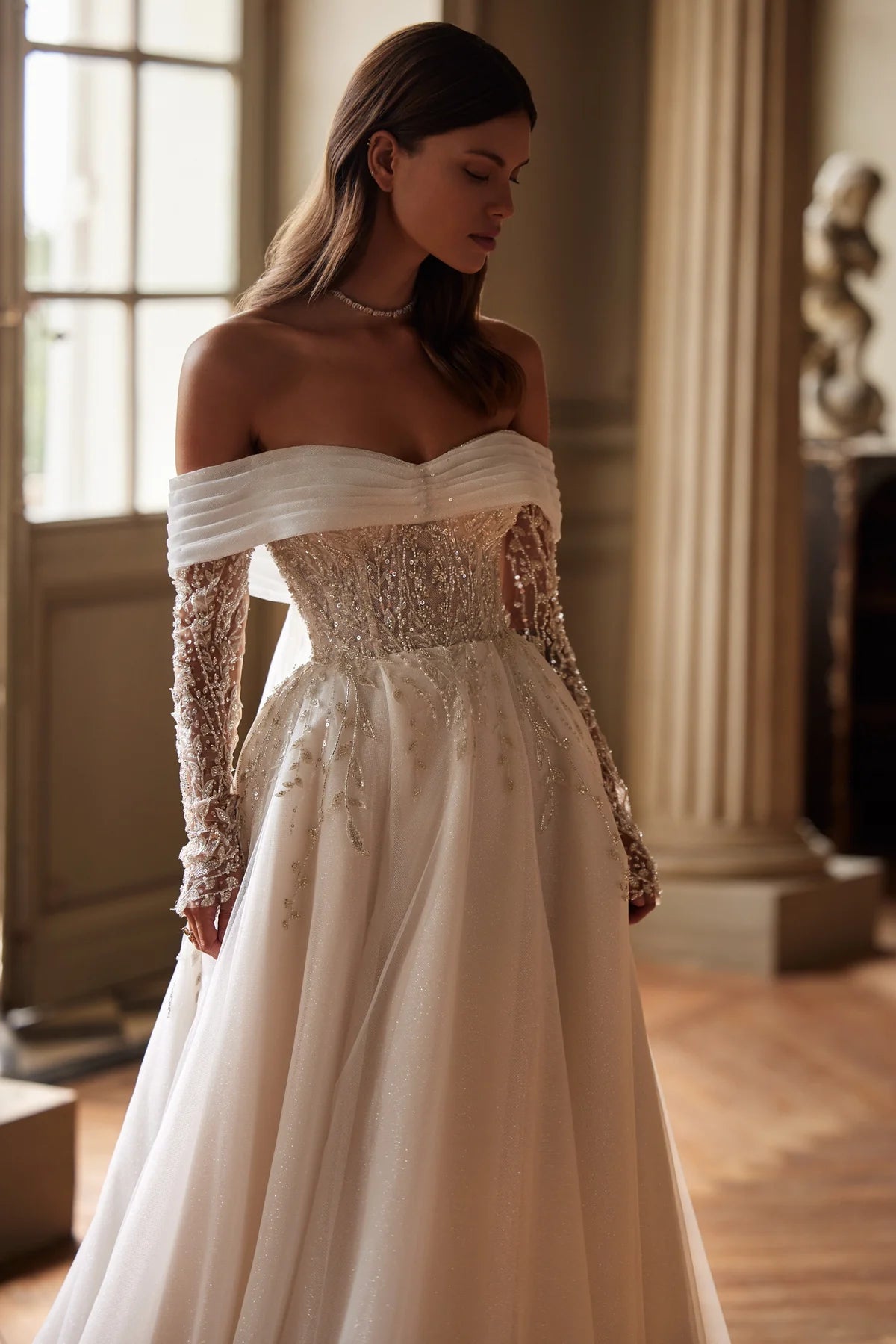 Indulge in pure elegance with the Josette Gown. A timeless A-line silhouette in ivory tone, crafted from sparkling tulle. The revealing neckline and long sleeves add a touch of sophistication. Adorned with silver lace embroidery, sequins, beads, pearls, and crystals. Tie the back zipper for a flawless fit.