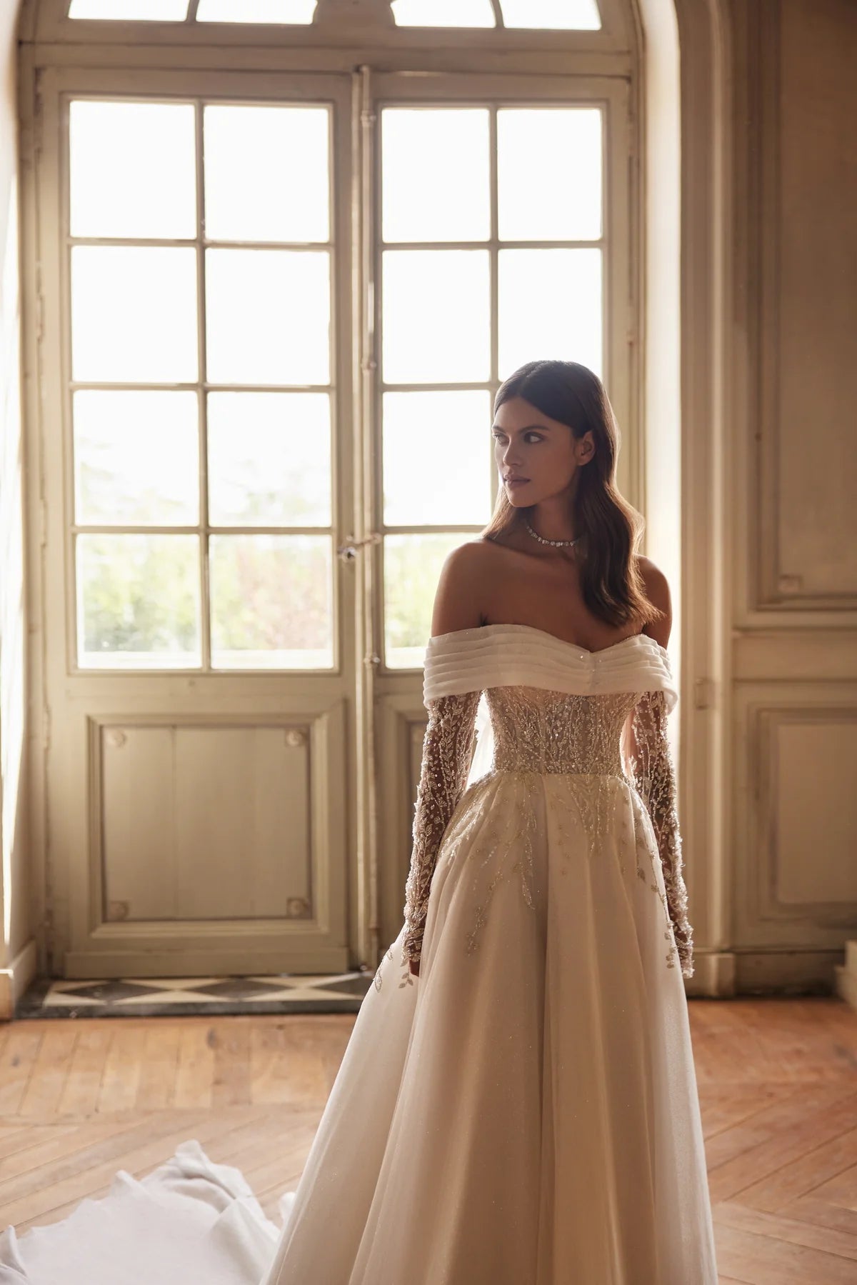 Indulge in pure elegance with the Josette Gown. A timeless A-line silhouette in ivory tone, crafted from sparkling tulle. The revealing neckline and long sleeves add a touch of sophistication. Adorned with silver lace embroidery, sequins, beads, pearls, and crystals. Tie the back zipper for a flawless fit.