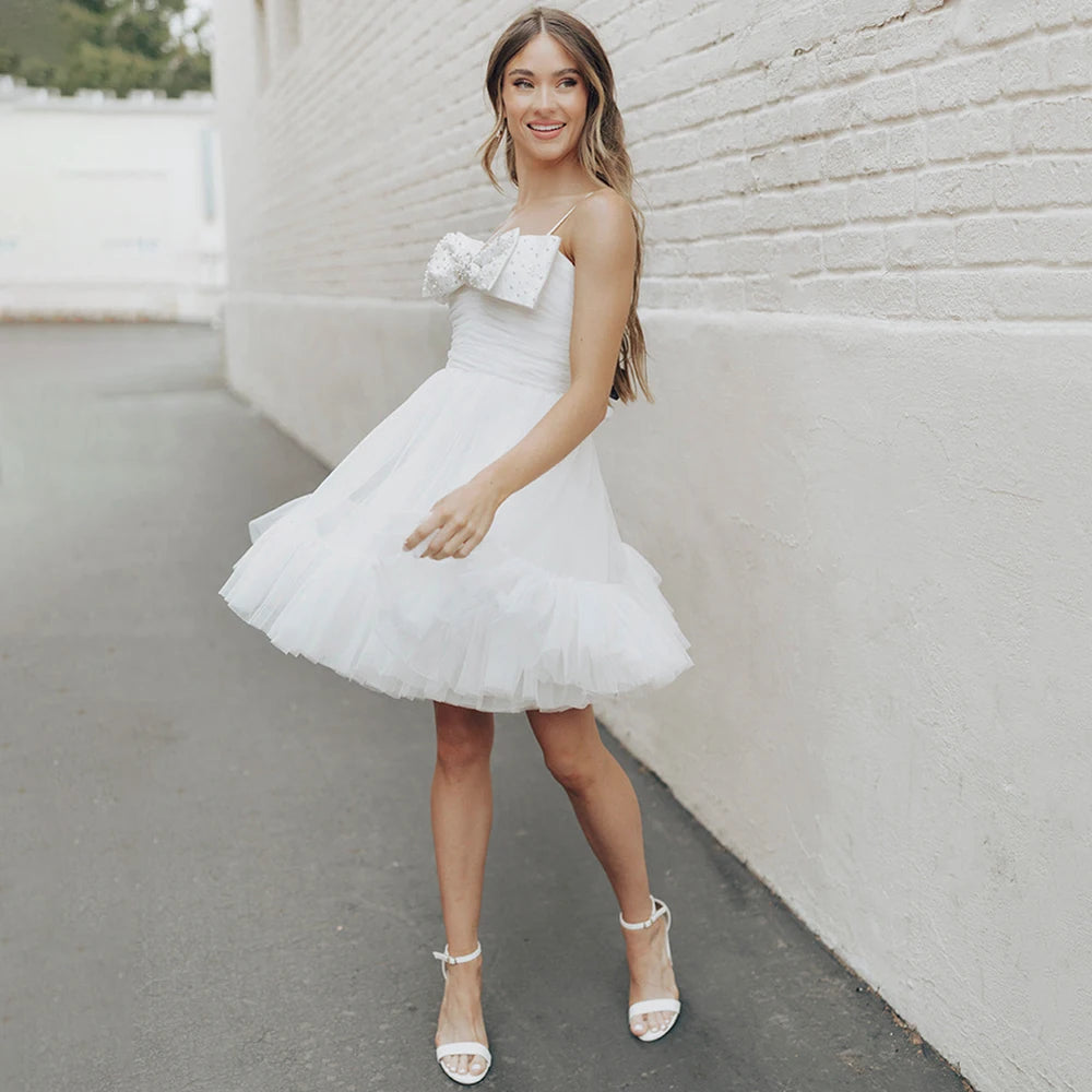 Experience the beauty and elegance of our Dress Elaina. With its beading detail, bow accents, and delicate tulle fabric, this wedding gown is perfect for a simple and charming look. The open back and spaghetti straps add a touch of playfulness, while the pearls and princess silhouette bring a sense of refinement. Say "I do" in style with this above knee bridal gown.