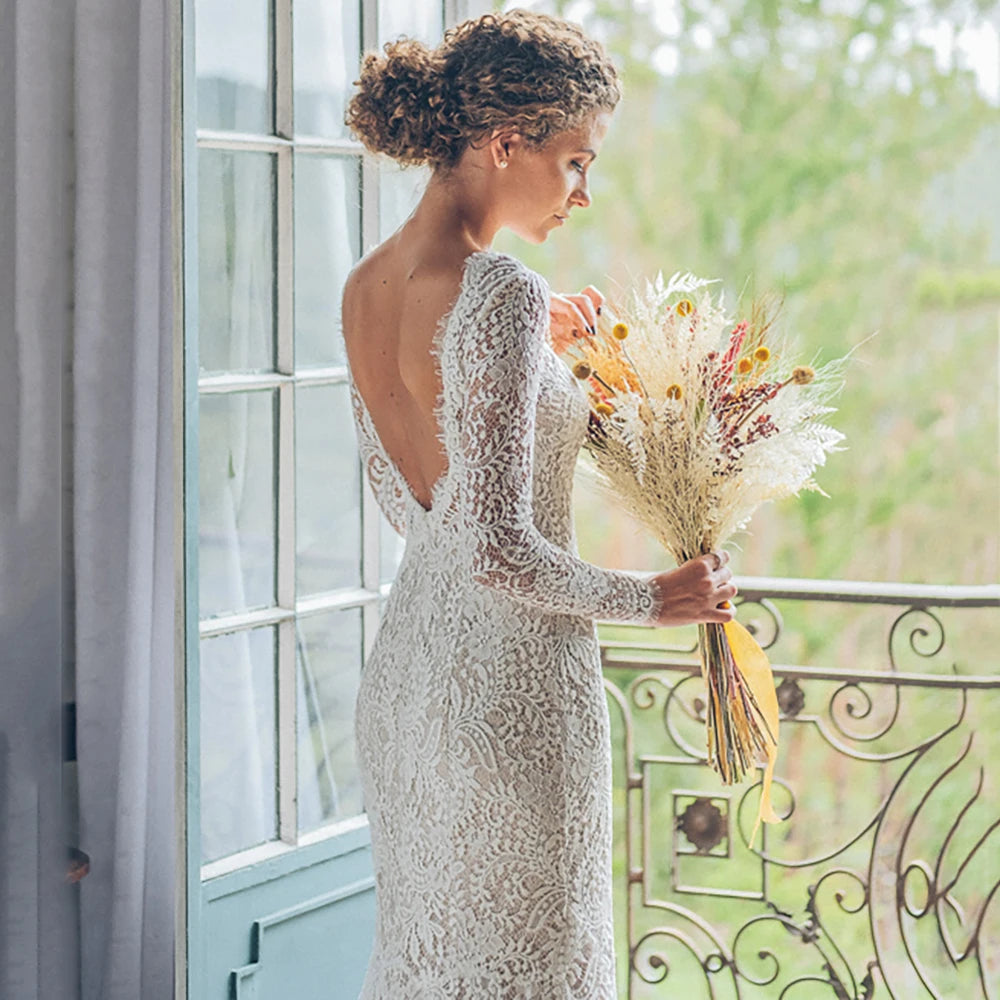Look breathtaking in the Eleanore, a romantic boho wedding gown crafted in allover lace. Perfect for a graceful mermaid silhouette, this custom-made gown features long sleeves, a boat neckline, and an open back for a truly captivating look. The slim fit flatters the figure, while delicate eyelash lace detailing adds to the classic, yet modern style. An enchanting choice for your special day.