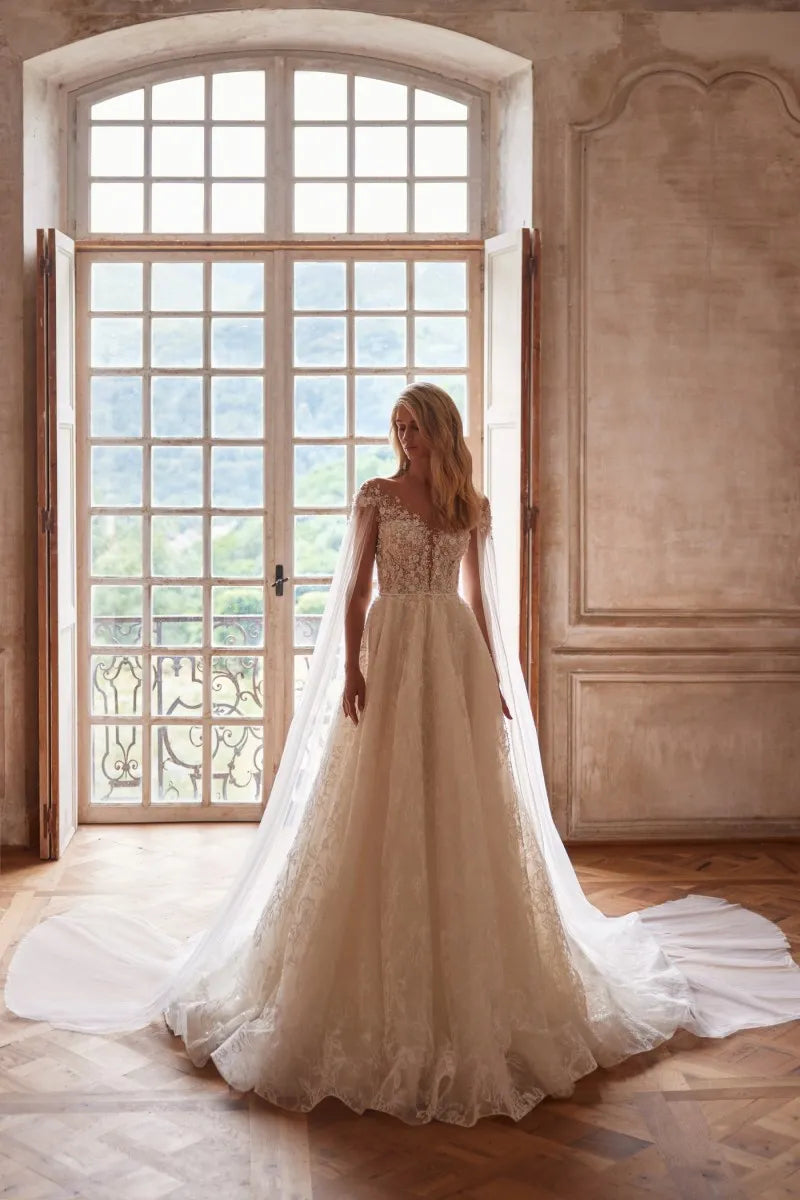 Elevate your wedding day look with our Dress Chantalle! Featuring a classic A-line silhouette, scoop neckline, and detachable cape sleeves, this dress is the epitome of elegance. Delicate appliques lace and a sophisticated train add romantic details to this modern bride gown. Make your special day unforgettable with Robe Chantalle.