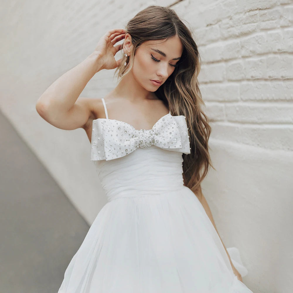 Experience the beauty and elegance of our Dress Elaina. With its beading detail, bow accents, and delicate tulle fabric, this wedding gown is perfect for a simple and charming look. The open back and spaghetti straps add a touch of playfulness, while the pearls and princess silhouette bring a sense of refinement. Say "I do" in style with this above knee bridal gown.