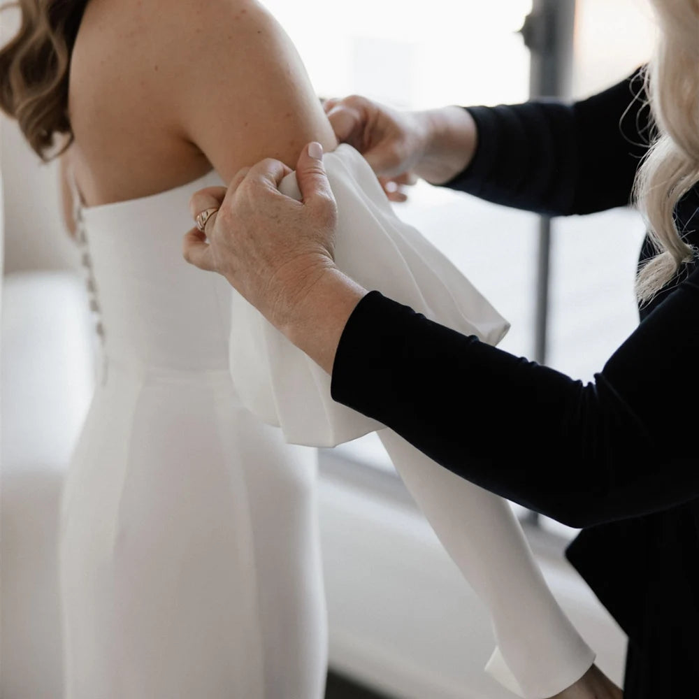Introducing the Robe Eglantine, a minimalist wedding dress that exudes effortless elegance. This ivory crepe gown features detachable long sleeves for versatility and a sleek sheath silhouette with a brush train for a modern touch. The strapless design and buttons closure add convenience for the bride.