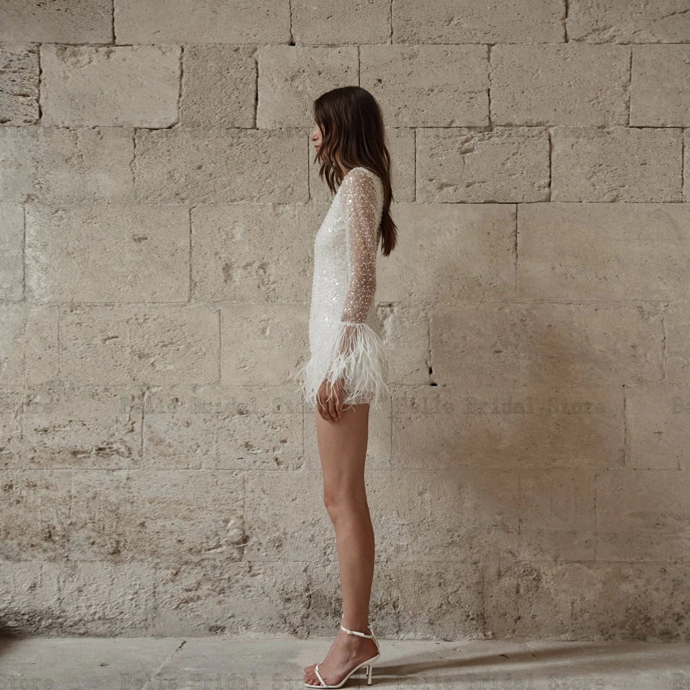Sparkle on your special day with our Robe Chantal. This glittery short wedding dress features an O neck, long sleeves, and a playful mini length with feather details. Guaranteed to make a statement and turn heads as you walk down the aisle.