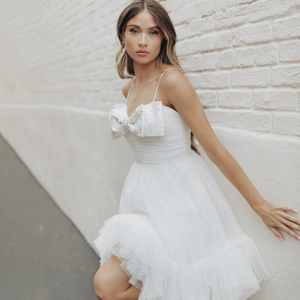 Experience the beauty and elegance of our Dress Elaina. With its beading detail, bow accents, and delicate tulle fabric, this wedding gown is perfect for a simple and charming look. The open back and spaghetti straps add a touch of playfulness, while the pearls and princess silhouette bring a sense of refinement. Say "I do" in style with this above knee bridal gown.