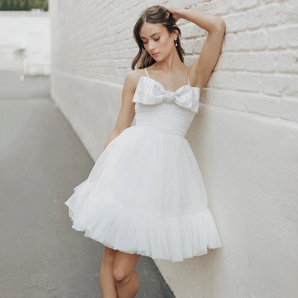 Experience the beauty and elegance of our Dress Elaina. With its beading detail, bow accents, and delicate tulle fabric, this wedding gown is perfect for a simple and charming look. The open back and spaghetti straps add a touch of playfulness, while the pearls and princess silhouette bring a sense of refinement. Say "I do" in style with this above knee bridal gown.