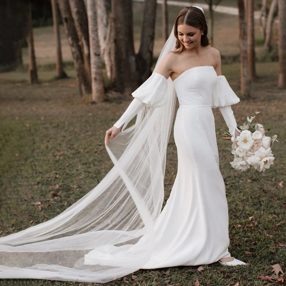 Introducing the Robe Eglantine, a minimalist wedding dress that exudes effortless elegance. This ivory crepe gown features detachable long sleeves for versatility and a sleek sheath silhouette with a brush train for a modern touch. The strapless design and buttons closure add convenience for the bride.