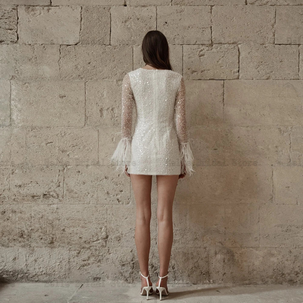Sparkle on your special day with our Robe Chantal. This glittery short wedding dress features an O neck, long sleeves, and a playful mini length with feather details. Guaranteed to make a statement and turn heads as you walk down the aisle.