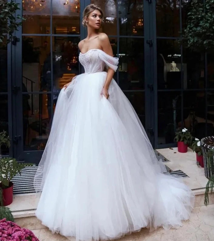 Elevate your wedding day look with our Robe Melina. This elegant ball gown boasts intricate beading, an off the shoulder sweetheart neckline, and a sleeveless tulle design. Perfect for the sophisticated bride seeking a touch of luxury and glamour.