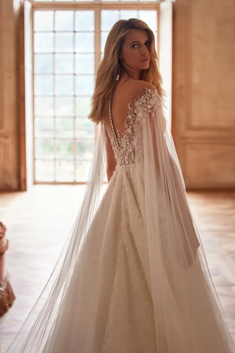 Elevate your wedding day look with our Dress Chantalle! Featuring a classic A-line silhouette, scoop neckline, and detachable cape sleeves, this dress is the epitome of elegance. Delicate appliques lace and a sophisticated train add romantic details to this modern bride gown. Make your special day unforgettable with Robe Chantalle.