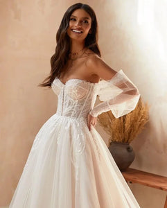 Elevate your bridal style with the Robe Vittoria. This charming A-line dress features a sweetheart neckline, intricate lace appliques, and long puff sleeves with delicate ruffles. Perfect for a boho-inspired wedding, this dress exudes elegance and romance. Say "I do" in style with the Robe Vittoria.
