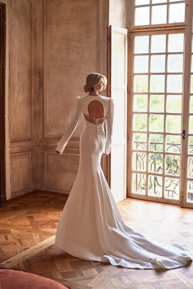 Elevate your wedding day look with the versatile Robe Coquette. This 2 in 1 dress features a detachable skirt, allowing you to easily transition from a stunning mermaid silhouette to a sexy high split. The long sleeves and sweetheart neckline are adorned with beautiful beadwork, adding a touch of elegance to the crepe fabric. Perfect for the modern bride.