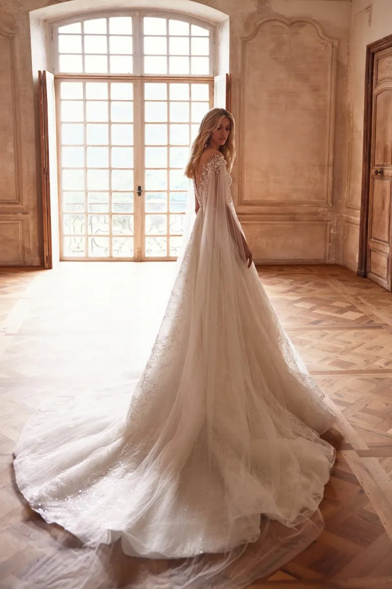 Elevate your wedding day look with our Dress Chantalle! Featuring a classic A-line silhouette, scoop neckline, and detachable cape sleeves, this dress is the epitome of elegance. Delicate appliques lace and a sophisticated train add romantic details to this modern bride gown. Make your special day unforgettable with Robe Chantalle.