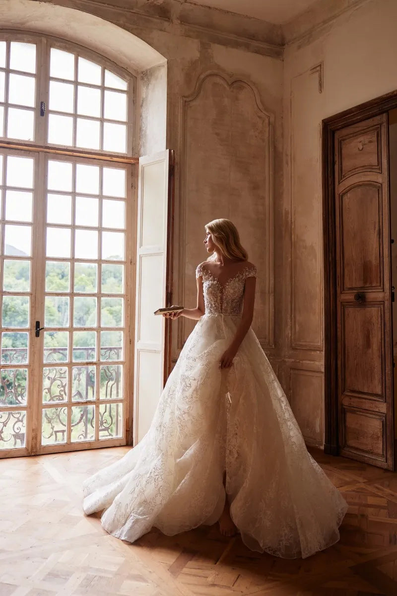 Elevate your wedding day look with our Dress Chantalle! Featuring a classic A-line silhouette, scoop neckline, and detachable cape sleeves, this dress is the epitome of elegance. Delicate appliques lace and a sophisticated train add romantic details to this modern bride gown. Make your special day unforgettable with Robe Chantalle.