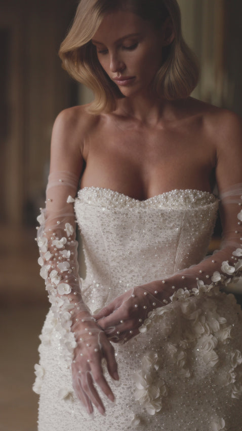 A dazzling bustier mini dress embellished with sequins, pearls, and 3D flowers by hand. Featuring a heart-shaped neckline with accentuated built-in cups, a cinched waist, and a structured tulip skirt adorned with 3D tulle flowers that add a sculpted finish. The look can be elevated with tulle long gloves adorned with volumetric flower appliqués. Perfect as a second wedding dress or a go-out party dress.